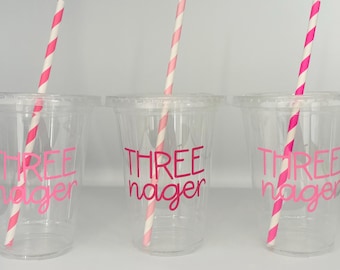 May include: Three clear plastic cups with pink text and pink and white striped straws. The text on the cups reads "THREE" over "nager". The cups are likely for a birthday celebration.