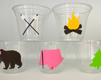 May include: Five clear plastic cups with different camping-themed designs. The designs include a campfire, a tent, a bear, a pair of crossed sticks, and a pine tree.