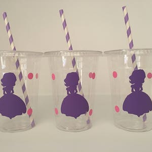 May include: Three clear plastic cups with purple and pink designs. Each cup has a purple silhouette of a princess with a crown and a pink polka dot pattern. Each cup has a purple and white striped straw.
