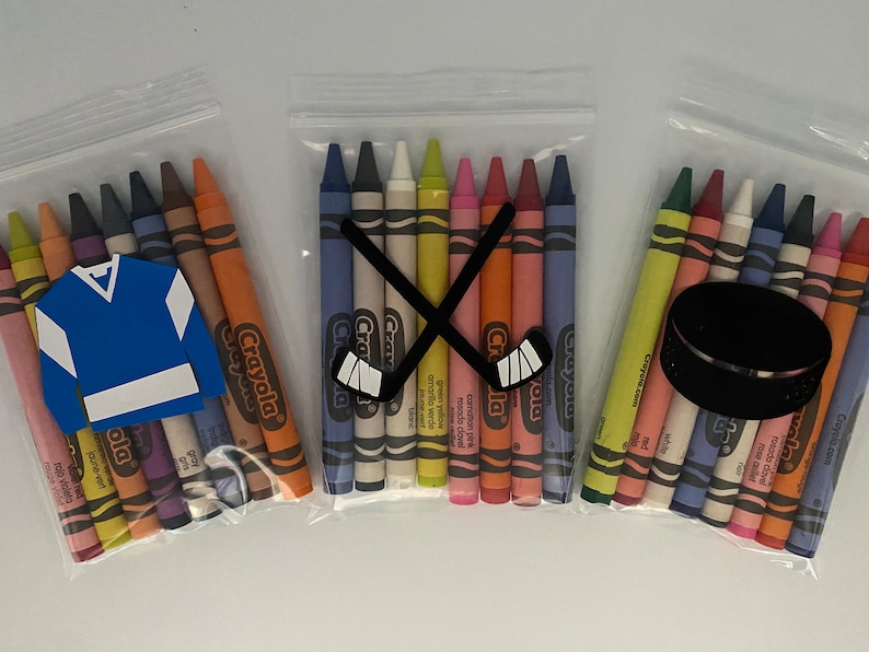 May include: Three clear plastic bags filled with Crayola crayons. The bags are decorated with hockey-themed cutouts. One bag has a blue hockey jersey, one has a pair of crossed hockey sticks, and one has a black hockey puck.