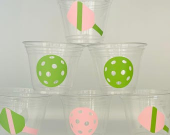 May include: Six clear plastic cups with green and pink designs. The designs include a pickleball paddle and a pickleball.