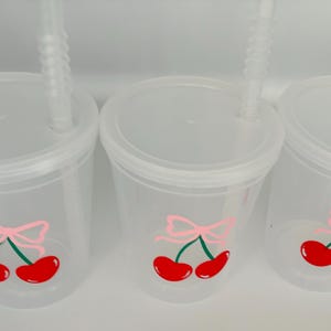 May include: Three clear plastic cups with lids and straws. Each cup has a pink bow and two red cherries with green stems drawn on the side.