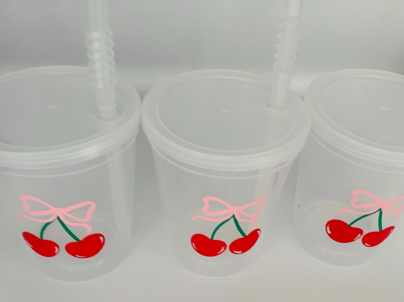 May include: Three clear plastic cups with lids and straws. Each cup has a pink bow and two red cherries with green stems drawn on the side.