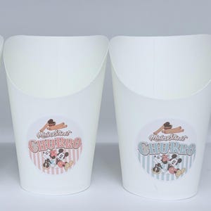 Op de afbeelding: Twee witte papieren voedselcontainers met ronde stickers. De stickers tonen de tekst Main Street CHURRO, een illustratie van churros en een afbeelding van twee tekenfilmmuizen. De ene sticker heeft een roze gestreepte achtergrond, de andere blauw.
