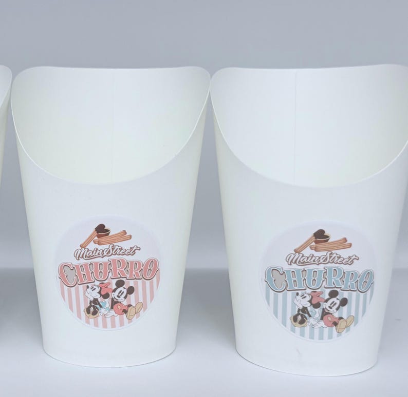 Op de afbeelding: Twee witte papieren voedselcontainers met ronde stickers. De stickers tonen de tekst Main Street CHURRO, een illustratie van churros en een afbeelding van twee tekenfilmmuizen. De ene sticker heeft een roze gestreepte achtergrond, de andere blauw.