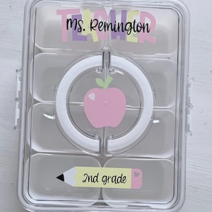 May include: A clear plastic lunchbox with multiple compartments. The lid features the text "TEACHER Ms. Remington" in pastel colors. A pink apple graphic is in the center, and a sticker that says "2nd grade" is at the bottom.