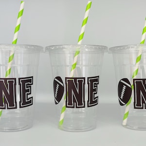 May include: Three clear plastic cups with lids, each featuring a brown football graphic and the word "ONE" in a bold font. Green and white striped straws are inserted into each cup. The cups are designed for a football-themed event.