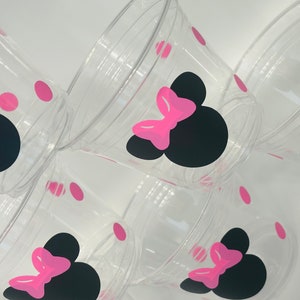Puede incluir: Conjunto de seis vasos de pl&aacute;stico transparente con dise&ntilde;os de Minnie Mouse en negro y rosa. Cada vaso tiene una silueta negra de la cabeza de Minnie Mouse con un lazo rosa y lunares rosas.