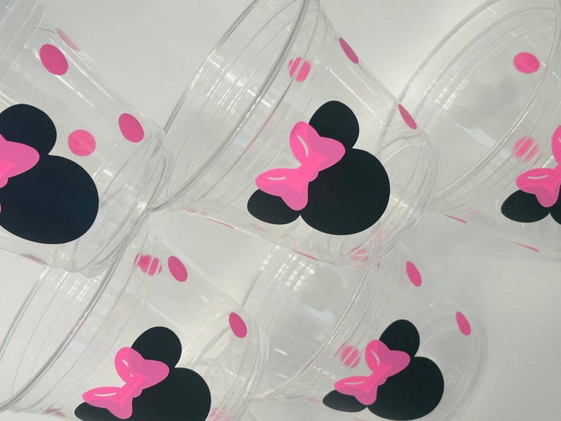 Puede incluir: Conjunto de seis vasos de pl&aacute;stico transparente con dise&ntilde;os de Minnie Mouse en negro y rosa. Cada vaso tiene una silueta negra de la cabeza de Minnie Mouse con un lazo rosa y lunares rosas.