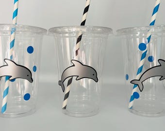 Puede incluir: Tres vasos de plástico transparentes con pajitas rayadas azules y blancas. Cada vaso tiene una pegatina de delfín con lunares azules.
