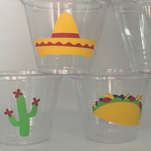 May include: Clear plastic cups with festive Mexican-themed designs. The cups feature a yellow sombrero, a green cactus with red flowers, and a yellow taco with green lettuce and red tomato.