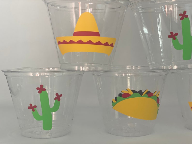 May include: Clear plastic cups with festive Mexican-themed designs. The cups feature a yellow sombrero, a green cactus with red flowers, and a yellow taco with green lettuce and red tomato.