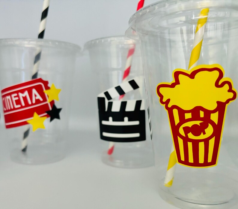 May include: Three clear plastic cups with red and white striped straws. Each cup has a different movie-themed sticker: one says "CINEMA" with a red ticket and stars, one has a black and white film clapboard, and one has a yellow popcorn bucket with red stripes.