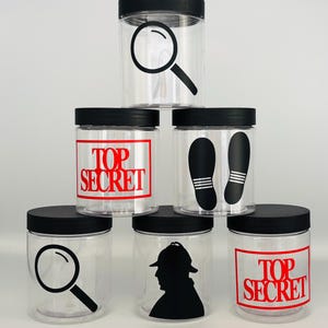 May include: Six clear glass jars with black lids. Each jar has a different black and white design on the front. The designs include a magnifying glass, footprints, a silhouette of a detective, and the words "Top Secret".