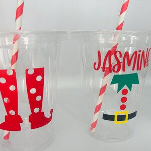 May include: Two clear plastic cups with red and white striped straws. One cup has a red and white elf boot design and the other has a green elf hat and the name "Jasmine" in red.