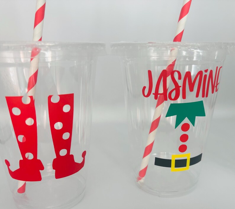 May include: Two clear plastic cups with red and white striped straws. One cup has a red and white elf boot design and the other has a green elf hat and the name "Jasmine" in red.