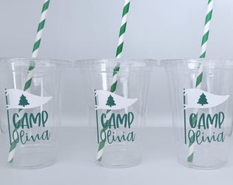 May include: Three clear plastic cups with lids and green and white striped straws. Each cup has a white flag design with a green tree and the words "CAMP Olivia" in green.