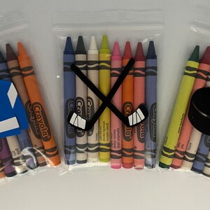 May include: Three bags of Crayola crayons with hockey-themed decorations. The first bag has a blue and white hockey jersey, the second bag has two crossed hockey sticks, and the third bag has a black hockey puck.