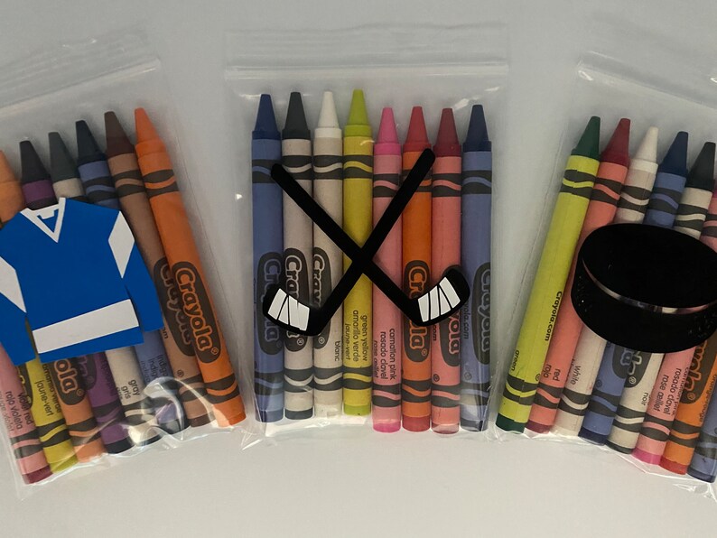 May include: Three bags of Crayola crayons with hockey-themed decorations. The first bag has a blue and white hockey jersey, the second bag has two crossed hockey sticks, and the third bag has a black hockey puck.
