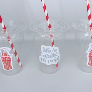 May include: Three clear plastic cups with lids and red and white striped straws. Each cup has a sticker with a Coca-Cola bottle design or the text "She's ready to pop!" in a light blue color.