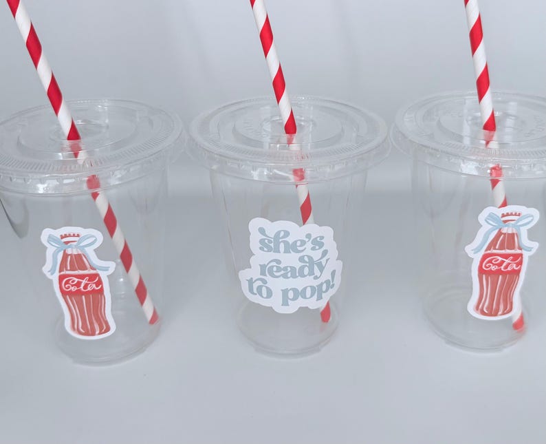 May include: Three clear plastic cups with lids and red and white striped straws. Each cup has a sticker with a Coca-Cola bottle design or the text "She's ready to pop!" in a light blue color.