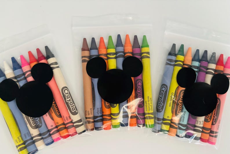 May include: Three clear plastic bags each filled with a set of Crayola crayons. Each bag has a black Mickey Mouse head shape attached. The crayons are various colors, including red, pink, blue, and yellow.