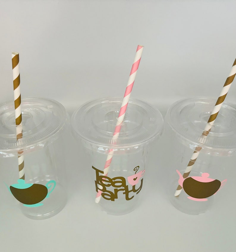 Puede incluir: Tres vasos de pl&aacute;stico transparente con tapas y pajitas de papel a rayas. Cada vaso tiene un dise&ntilde;o diferente con tem&aacute;tica de t&eacute;: una tetera, una taza de t&eacute; y las palabras "Tea Party". Las pajitas son doradas y blancas, y rosas y blancas.