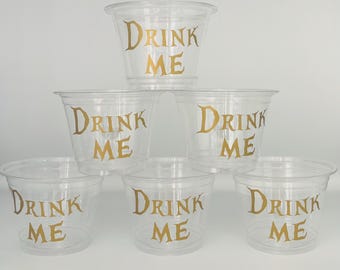 May include: Seven clear plastic cups stacked in a pyramid formation. Each cup has the words "DRINK ME" printed in gold. The cups are made of clear plastic and are suitable for a party or event.