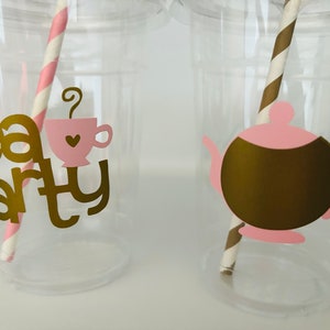 Puede incluir: Vasos de pl&aacute;stico transparente con texto dorado "Tea Party". Uno tiene un gr&aacute;fico de taza de t&eacute; rosa, el otro una tetera dorada. Cada vaso tiene una pajita de papel a rayas en rosa y blanco o marr&oacute;n y dorado.
