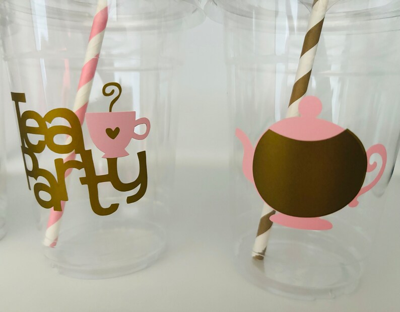 Puede incluir: Vasos de pl&aacute;stico transparente con texto dorado "Tea Party". Uno tiene un gr&aacute;fico de taza de t&eacute; rosa, el otro una tetera dorada. Cada vaso tiene una pajita de papel a rayas en rosa y blanco o marr&oacute;n y dorado.