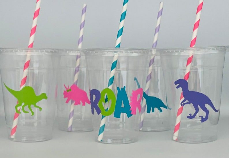 May include: Clear plastic cups with colorful dinosaur silhouettes and striped straws. The word "ROAR" is displayed on one cup. The dinosaurs are in green, pink, teal, and purple. The straws have pink, blue, and purple stripes.