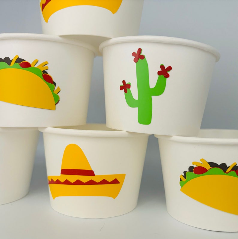 May include: A stack of six white paper cups with different Mexican-themed designs. The designs include a yellow sombrero, a green cactus with red flowers, and a yellow taco.