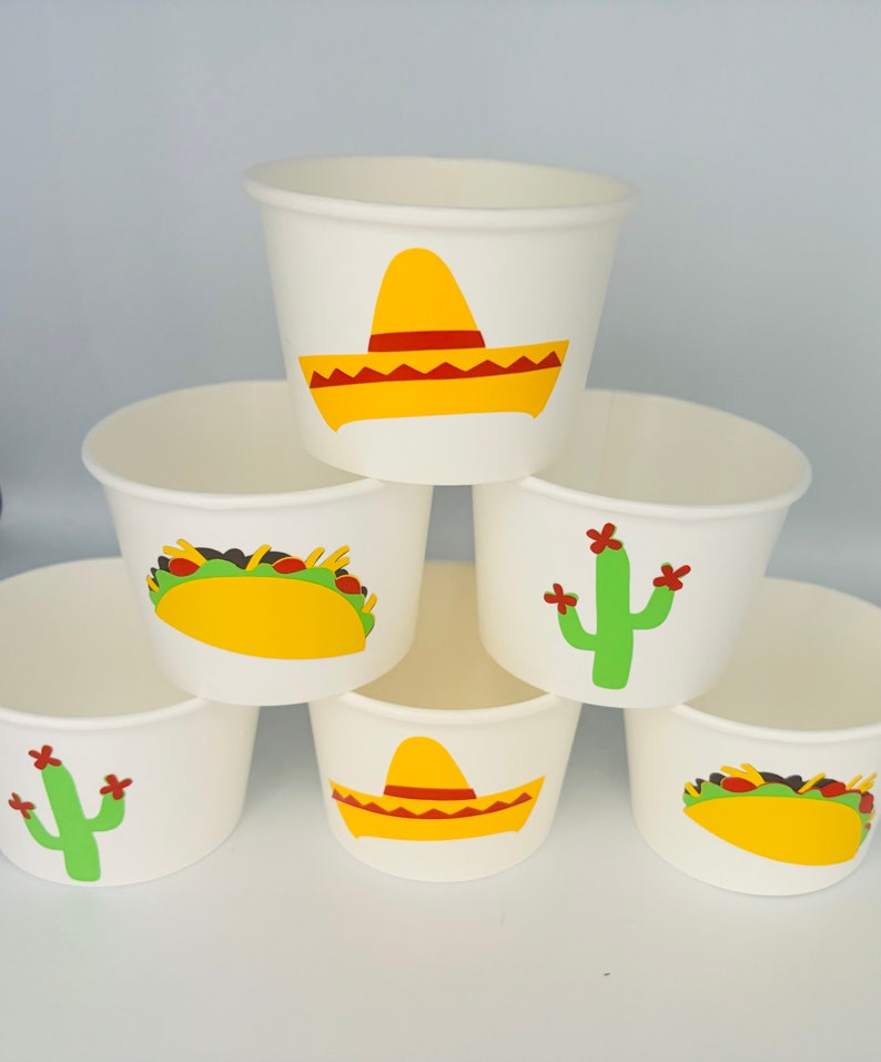 May include: Six white paper cups with different Mexican-themed designs. The designs include a sombrero, a cactus, and a taco.