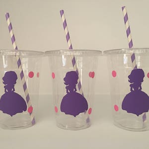 May include: Three clear plastic cups with purple and white striped straws. Each cup has a pink polka dot pattern and a purple silhouette of a princess wearing a dress.