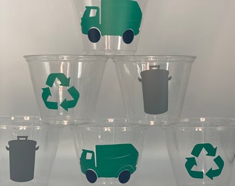 May include: Six clear plastic cups with green and gray recycling themed designs. The designs include a recycling symbol, a garbage can, and a green garbage truck.