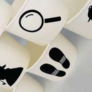 Puede incluir: Conjunto de seis vasos de papel blanco con dise&ntilde;os de silueta negra. Los dise&ntilde;os incluyen una lupa, huellas y una silueta de una persona con sombrero.