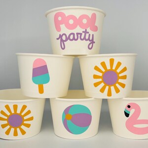 May include: Six white paper cups with different summer-themed designs. The cups have a pink ice cream cone, a yellow sun, a purple and green beach ball, a pink flamingo, and two yellow suns. The top cup says "Pool party" in pink and purple letters.