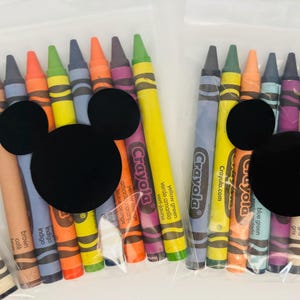 May include: Two clear plastic bags filled with Crayola crayons in various colors. Each bag features a black Mickey Mouse head shape. The crayons are labeled with their respective colors, including brown, indigo, and yellow green.