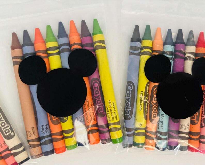 May include: Two clear plastic bags filled with Crayola crayons in various colors. Each bag features a black Mickey Mouse head shape. The crayons are labeled with their respective colors, including brown, indigo, and yellow green.