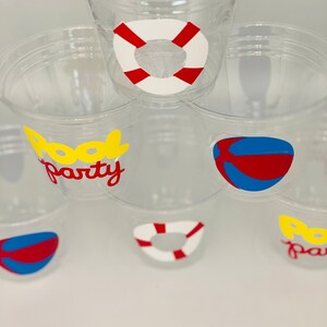May include: Clear plastic cups with pool party themed stickers. The stickers feature a red and white life preserver, a blue and red beach ball, and the words "Pool Party" in yellow and red letters.