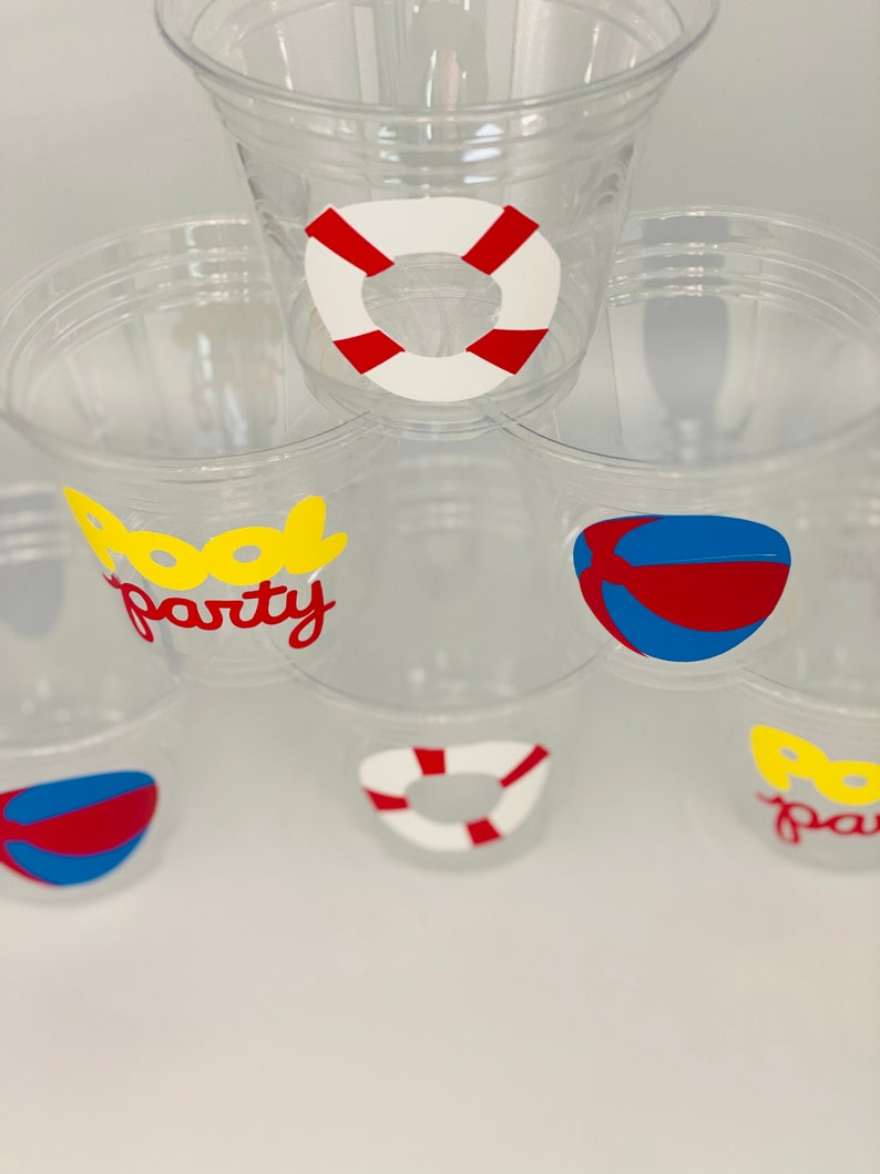 May include: Clear plastic cups with pool party themed stickers. The stickers feature a red and white life preserver, a blue and red beach ball, and the words "Pool Party" in yellow and red letters.