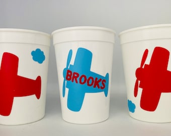 May include: Three white plastic cups with red and blue airplane designs. The middle cup has the name "BROOKS" written in red on the airplane.