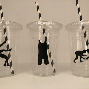 K&ouml;nnte beinhalten: Drei durchsichtige Plastikbecher mit schwarz-wei&szlig; gestreiften Strohhalmen. Jeder Becher hat eine schwarze Silhouette eines Wrestlers darauf. Ein Becher zeigt einen Wrestler in einem Wrestling-Tanktop, ein anderer einen Wrestler, der einen Salto macht, und der letzte zwei Wrestler, die ringen.