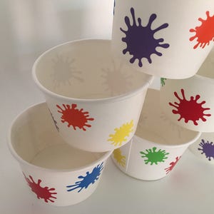 K&ouml;nnte beinhalten: Stapel aus sechs wei&szlig;en Pappbechern mit bunten Farbklecksmustern. Die Becher sind pyramidenf&ouml;rmig gestapelt, wobei der oberste Becher einen lilafarbenen Klecks zeigt, der n&auml;chste Becher einen roten Klecks und der unterste Becher einen roten, blauen und gelben Klecks.