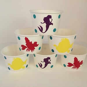 May include: Six white paper cups with teal polka dots and various colourful cartoon characters, including a mermaid, a crab, and a fish.