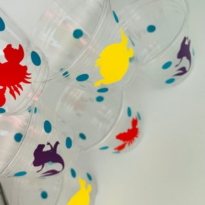 Puede incluir: Vasos de pl&aacute;stico transparentes con lunares azules y siluetas de personajes coloridos. Los personajes incluyen una langosta roja, un pez amarillo, una sirena morada y un pez naranja.