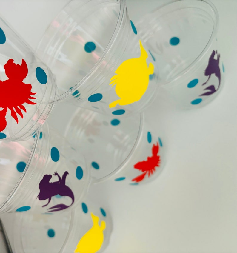 Puede incluir: Vasos de pl&aacute;stico transparentes con lunares azules y siluetas de personajes coloridos. Los personajes incluyen una langosta roja, un pez amarillo, una sirena morada y un pez naranja.