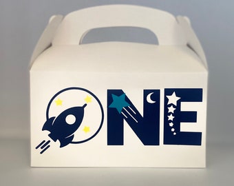 May include: White party favor box with a handle, decorated with a navy blue rocket ship, yellow stars, and the word "ONE" in navy blue with a shooting star and crescent moon. Perfect for a space-themed birthday party.