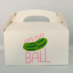 May include: White cardboard box with a handle. The box has a green pickle illustration and the text "Let's Play Ball" in pink.