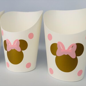 K&ouml;nnte beinhalten: Drei wei&szlig;e Pappbecher mit einer goldenen Minnie Mouse-Silhouette und einer rosa Schleife. Die Becher haben rosa Punkte.